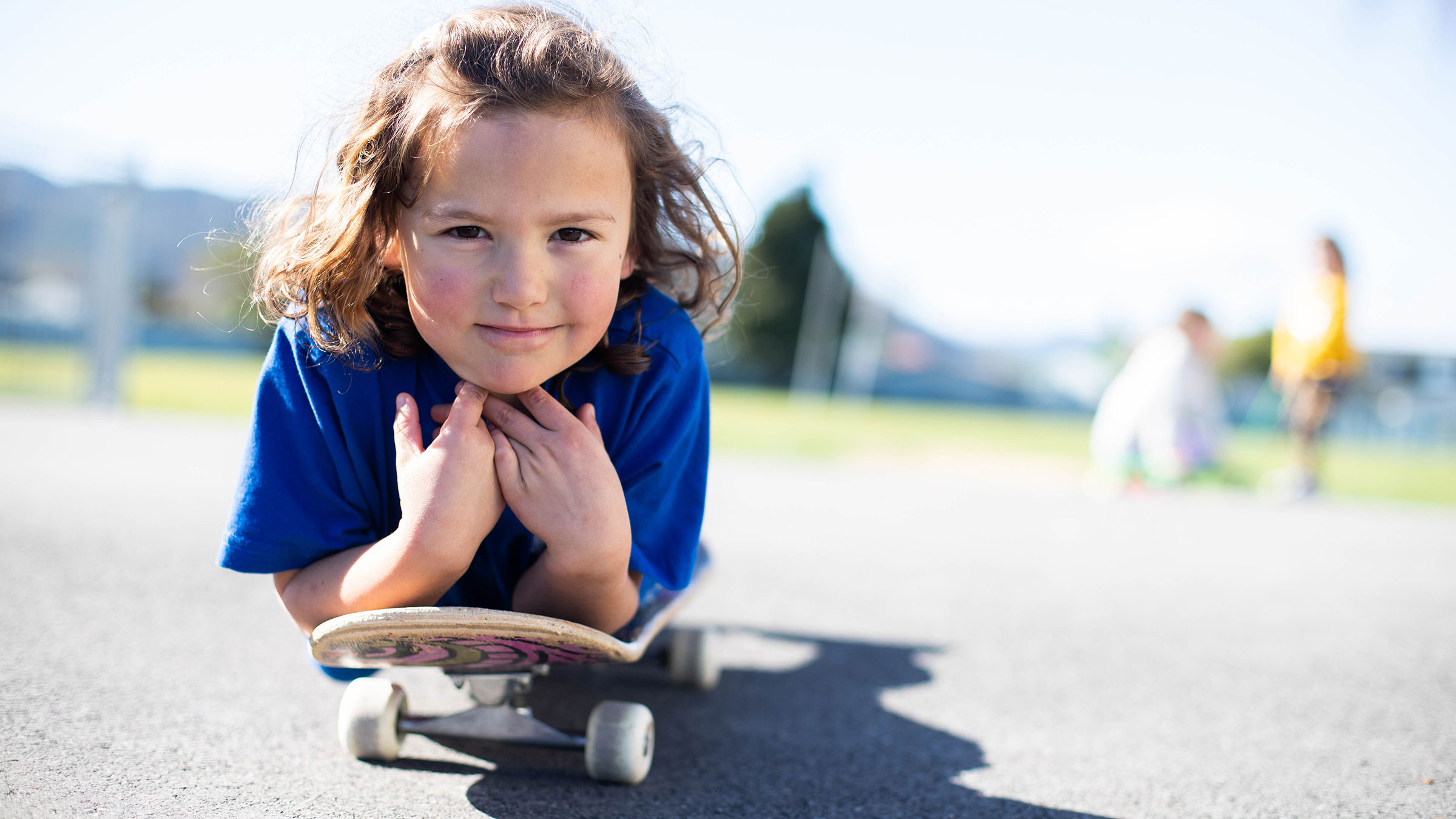 A student outdoors, lying on a skateboard and smiling